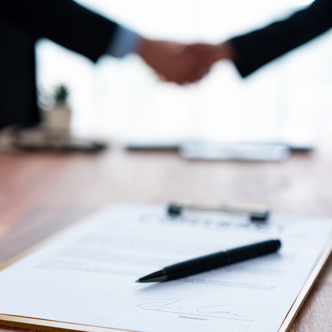 A close-up of a contract with a pen, while two business people shake hands in the background, symbolizing an agreement or partnership.